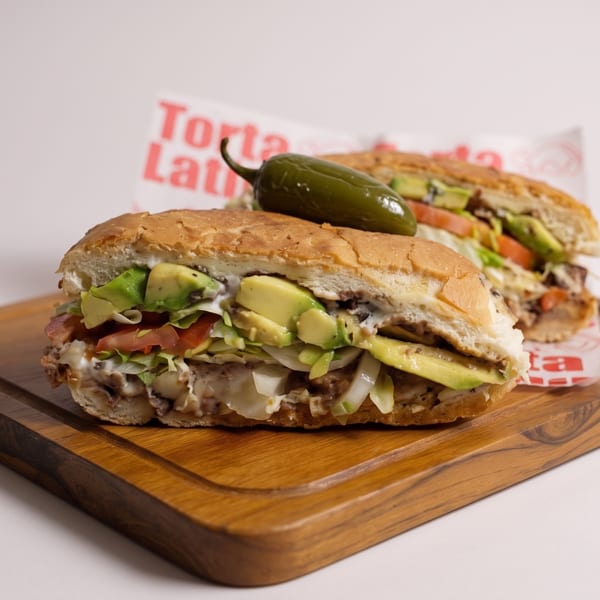 Two tortas, one with a jalapeño on top, filled with avocado, lettuce, tomato, and meat.