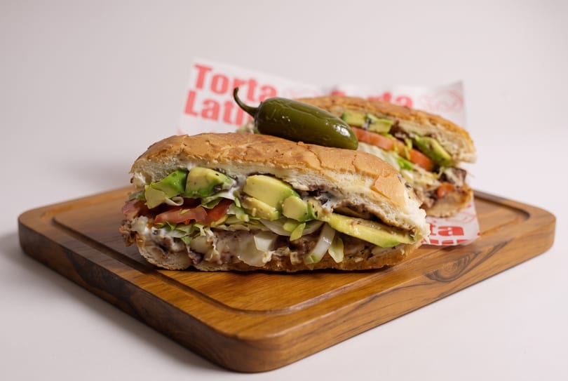 Two tortas, one with a jalapeño on top, filled with avocado, lettuce, tomato, and meat.